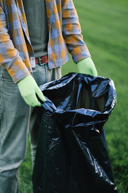 A person wearing a plaid shirt in shades of yellow, blue, and brown, with green gloves, is standing outdoors on a grassy area, holding open a black plastic rubbish bag with their left hand, preparing for rubbish collection. The individual’s trousers are grey with a brown belt. The background features a blurred green lawn or field, indicating an outdoor environment suitable for waste or garden refuse collection. The person is engaged in an activity related to waste removal, possibly part of an on-site clearance operation that might be handled by private waste removal services like Waste Removal Brent. This scene emphasizes the process of loading refuse into a bin liner, aligning with the themes of rubbish removal or alternative waste handling outside of local authority collection. The lighting is natural, and the focus is on the hands, bag, and the environment, providing a clear, factual representation of an independent rubbish collection activity involving waste management equipment and protective gloves, essential for hygiene and safety during rubbish handling.