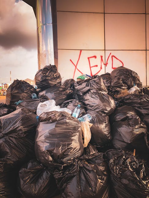 A large pile of black plastic rubbish bags filled with waste is stacked against a modern building with a tiled exterior wall, which is partially visible in the background. Some bags are slightly torn, revealing glimpses of their contents, and a few transparent plastic bottles and crumpled paper items are protruding from the bags. The surrounding area appears to be an outdoor urban setting, possibly a designated drop-off or temporary collection point, with a cloudy sky overhead. The scene is indicative of private waste collection and rubbish removal services, such as those provided by Waste Removal Brent, which handle on-site clearance outside traditional council refuse collection. The bags vary in size and are piled unevenly, creating a chaotic impression of waste waiting to be collected or disposed of. In the background, subtle hints of a city skyline can be seen, emphasizing the urban environment and the importance of managing waste responsibly. The lighting suggests late afternoon or early evening, with warm tones cast on the scene.