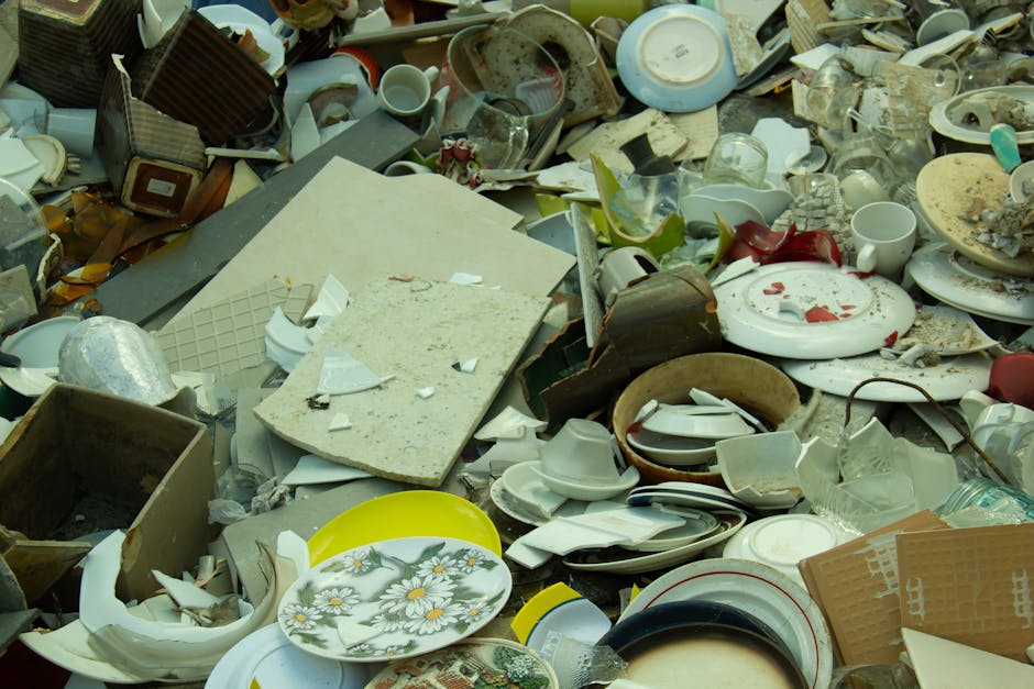The image displays a large pile of assorted broken and discarded household crockery, ceramics, and kitchenware, including plates, bowls, cups, and saucers, scattered across a surface, likely outdoors. The ceramics vary in color, with many predominantly white but some pieces showing floral or patterned designs in shades of green, yellow, and red. Several items are chipped or shattered, with fragments and debris spread throughout the heap. A rectangular piece of broken beige stone or tile with a textured surface lies amidst the collection. Surrounding the ceramics are various other materials such as cardboard boxes, glass shards, and plastic items, contributing to a sense of clutter typical of waste disposal scenes. The environment appears to be an outdoor setting, possibly a driveway or garden area, under natural daylight, highlighting the chaotic accumulation of household waste awaiting professional rubbish removal services. Waste Removal Brent’s presence in waste management is implied through the scene, illustrating the need for proper disposal or collection of such bulky and broken items, often handled via private or alternative waste handling methods rather than standard council collection.