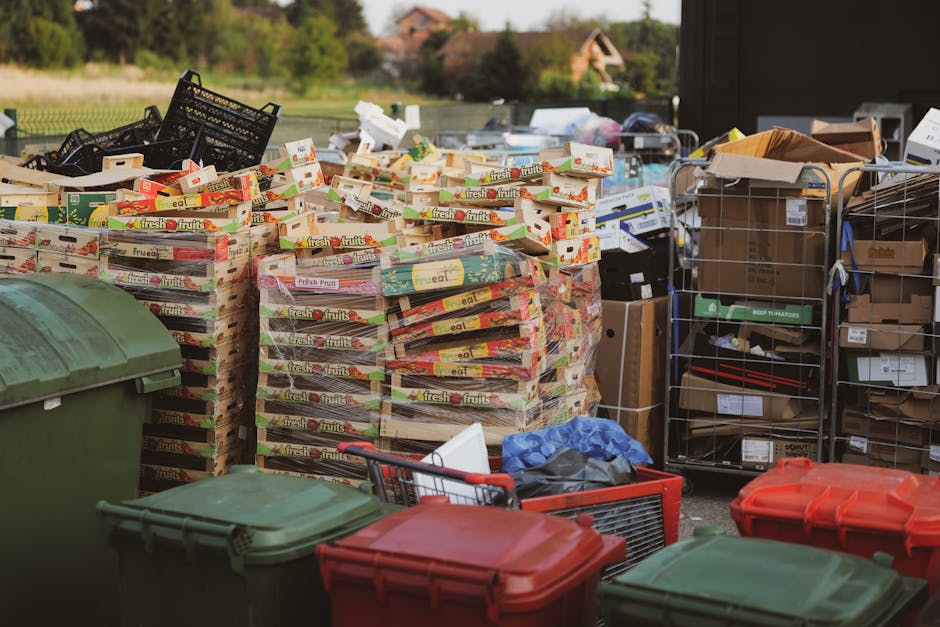 A densely packed outdoor scene featuring multiple stacks of cardboard boxes labeled 'fresh fruits' arranged on pallets, with some boxes slightly open revealing ajar packaging; the boxes are primarily in white, red, and green colors, with some appearing slightly crushed or weathered. To the left, a large green wheelie bin with a closed lid is visible, partially obscuring the lower part of the scene. On the right, there are metal wire cages filled with various flat-packed cardboard and paper-based materials, alongside open cardboard boxes containing additional waste items. In the foreground, red and green plastic wheelie bins with fitted lids are seen, some with lids slightly ajar or resting nearby, and a small shopping trolley with a black handle is placed among the bins and waste. The environment is an outdoor area, possibly a designated waste collection or sorting zone, with a paved surface and a background featuring greenery, trees, and residential houses under natural daylight. This scene illustrates a typical collection of bulky or recyclable waste that could be handled by private waste removal services similar to those offered by Waste Removal Brent, emphasizing the importance of proper sorting and disposal of such materials.