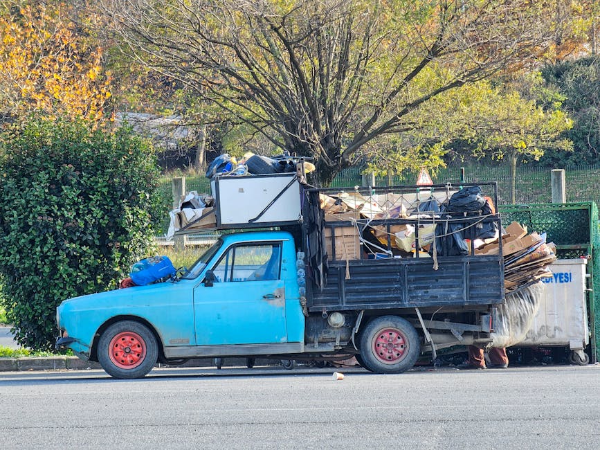 A small, aged blue flatbed truck is parked alongside a pavement, loaded with a large collection of assorted waste materials. The truck's body shows signs of wear, with faded paint and some rust visible on the metal frame, which is black and utilitarian in design. The front wheels are red and the body of the vehicle is compact, with a cab that has slightly cloudy windows. The flatbed area is filled to capacity with various debris, including cardboard boxes, black plastic bags, and loose waste, some tied with rope or string to secure the load. Behind the vehicle, a large tree with green and yellow leaves is visible, indicating an autumn setting, and a wooden fence runs parallel to the road in the background. The scene suggests an off-site, private collection potentially organized for rubbish removal or waste clearance services by Waste Removal Brent, highlighting an independent approach to rubbish disposal outside municipal services. The environment appears calm and clear, with natural daylight illuminating the scene.