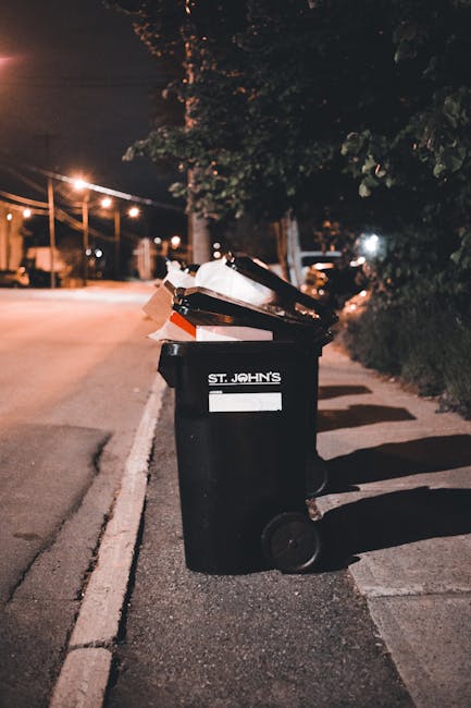 A black wheeled waste bin labeled 'St. John's' is positioned on the edge of a sidewalk on a quiet street at night. The lid of the bin is open, revealing various discarded items including cardboard boxes, paper, and plastic containers. The bin is made of durable plastic and has a textured surface. It stands upright on two wheels, with a small handle at the back. Shadows cast by streetlights stretch across the pavement, and in the background, there are faint outlines of trees, parked cars, and a row of streetlights illuminating the quiet street. The scene suggests routine rubbish collection, with the bin ready for waste removal services, possibly provided by a private waste collection company like Waste Removal Brent, which offers on-site clearance and alternative waste handling solutions.