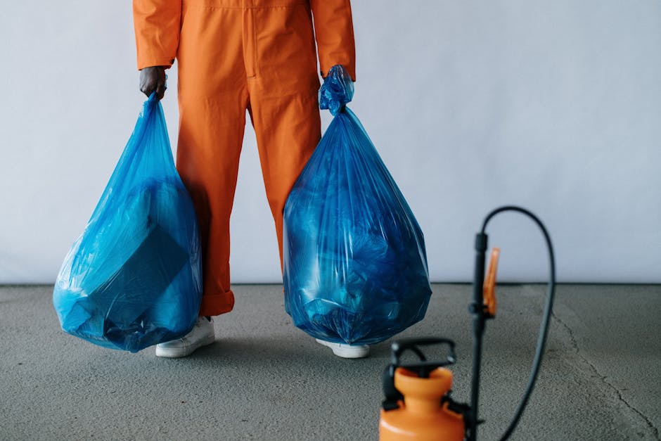 A person dressed in orange work overalls is holding two large blue plastic rubbish bags, each filled with waste, with one bag in each hand. The individual is standing on a bare, gray concrete floor in front of a plain, light-colored background. The bags are semi-transparent, showing the contents inside, and are tied at the top with the plastic gathered. Near the person's feet, a small portion of white footwear is visible. In the lower right corner of the image, there is an orange and black handheld sprayer or fogger, with a flexible tube attached, resting on the floor. This scene appears to depict an arrangement involving waste collection or on-site clearance, aligning with the services offered by Waste Removal Brent, especially for private, independent rubbish disposal or event clean-up activities where large waste bags are handled directly by personnel.
