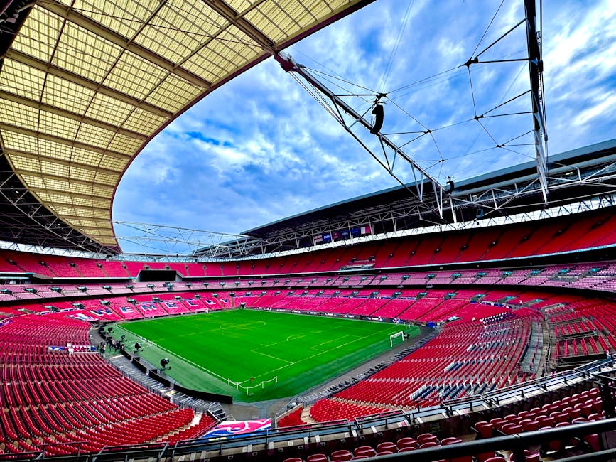 A large, empty sports stadium with a bright green grass pitch at the centre, surrounded by multiple tiers of red seating. The seating sections are arranged in curved rows that extend around the entire pitch. The stadium's roof structure arcs overhead, supported by black metal beams, with part of the roof covered in a beige, semi-transparent material that filters sunlight. Overhead, blue sky with scattered white clouds is visible through the open sections of the roof, and in the background, the stadium's upper stands and support structures are seen. The atmosphere appears bright and naturally lit, emphasizing the clean and well-maintained appearance of the venue. This scene illustrates a typical large-scale event space, relevant for considering private or alternative waste management options by Waste Removal Brent for event clean-ups or stadium waste clearance, with ample space for rubbish collection and disposal operations.
